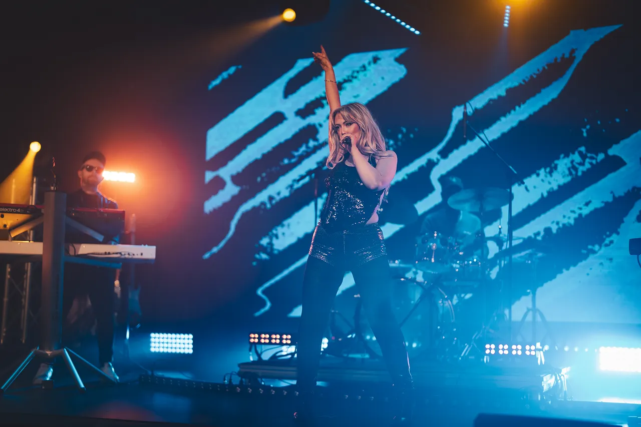 Female singer with long blonde hair performing on stage wearing a black sequined outfit and holding a microphone.